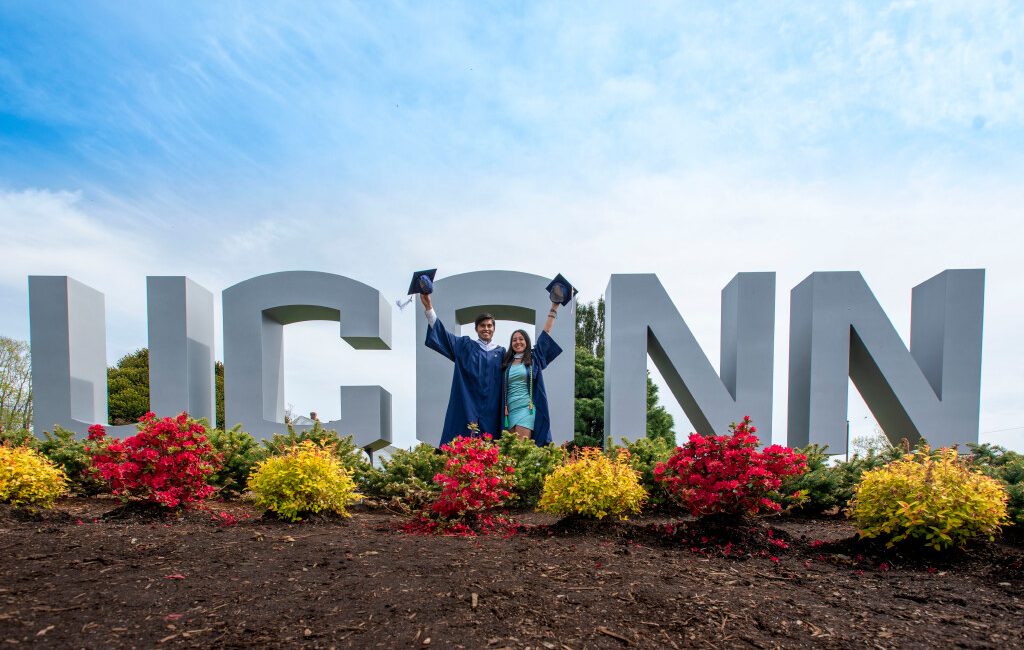 Two graduating seniors in caps and gowns in front of the UConn sign at the entrance to campus.